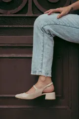 Zapato estilo Mary Jane con diseño destalonado, confeccionado en cuero color beige. Presenta una tira fina con hebilla metálica sobre el empeine, punta redondeada y taco cuadrado forrado de 6 cm de altura.