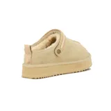 Pantufla tipo zueco color beige, confeccionada en cuero con costura central visible. Presenta una tira trasera ajustable con detalle tejido y suela de plataforma dentada.