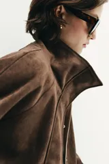 Chaqueta de tejido efecto ante color marrón, con cuello alto, cierre frontal con cremallera y botones a presión, manga larga y bolsillos de plastrón con solapa en el delantero.