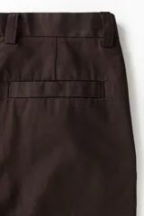 Pantalón sastrero color marrón oscuro, de corte relajado y piernas con pliegues, confeccionado en mezcla de algodón y lyocell. Cuenta con cierre y botón, bolsillos laterales y bolsillos traseros insertados con botón oculto.