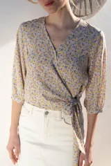 Blusa cruzada con estampado floral en tonos de lila, amarillo y verde. El cruce se ajusta a la cintura con un lazo. Tiene mangas 3/4 con puños elásticos y escote en V.