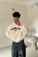 Sweater de punto color crudo con calce oversize y cuello redondo. Presenta bordados frontales en color negro con el texto "Scuffers", "Everyday Urban Aesthetics" y un pequeño logo circular. Confeccionado en una mezcla de acrílico, nylon, lana y spandex.