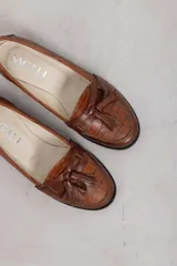 Mocasines de cuero marrón con diseño clásico, detalles de flecos y borlas en el empeine, y suela plana.