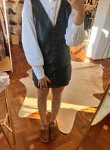 Vestido corto de cuero negro con escote en V, sin mangas y detalles de tachas metálicas a lo largo del frente y los bordes. Diseño recto con cierre frontal.