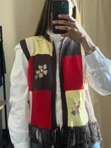 Chaleco tejido de punto con diseño de bloques de colores (marrón, rojo, amarillo y blanco), cuello redondo y detalles bordados de flores. Cuenta con flecos en el ruedo inferior.