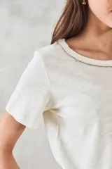 Remera color crudo de algodón peinado, de corte recto y mangas cortas. Presenta cuello redondo con detalle de cadena metálica dorada.