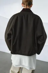 Chaqueta encerada de corte oversize, color marrón oscuro, con cuello de pana en contraste. Presenta cierre frontal con cremallera y botones a presión ocultos bajo solapa, bolsillos delanteros y mangas largas con puños ajustables.