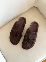 Zueco tipo mocasín de gamuza color marrón chocolate, con detalle de costuras visibles en el borde y cordones decorativos con borlas en el empeine.