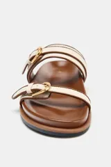 Sandalia plana color marrón con tiras de tela color crudo y hebillas doradas.