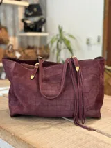 Cartera tipo tote de gamuza color bordó con textura de efecto cocodrilo. Presenta doble asa de hombro con detalles metálicos dorados y flecos largos decorativos en uno de los laterales.