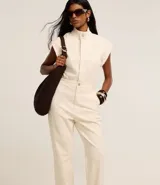 Mono largo de sarga color crudo, con cuello alto, cierre frontal con botones ocultos, sin mangas y diseño con trabillas en la cintura.