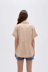 Camisa de viscosa color beige, con cuello camisero, mangas cortas, cierre frontal con botones ocultos y cordón ajustable en la cintura.
