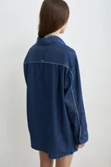 Camisa de jean azul, de corte holgado y manga larga, con cuello camisero y cierre frontal de botones oscuros.