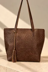 Bolso tipo tote bag color marrón chocolate, confeccionado 100% en cuero vacuno con textura símil cocodrilo. Presenta doble asa larga para hombro, costuras laterales con pasamanería y detalle colgante de borla de flecos de cuero.