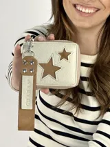 Billetera de cuero color crema con cierre perimetral y tira de cuero marrón con mosquetón y remaches. Presenta dos estrellas caladas en el frente, una grande y una pequeña.