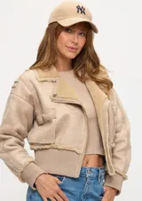 Campera corta tipo aviador en color beige, confeccionada en material con textura gamuzada y forro interno de corderito sintético. Presenta cierre frontal, bolsillos laterales con solapa y puños y ruedo en tejido acanalado.