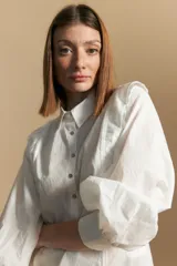 Camisa blanca de manga larga con cuello y botones frontales. Presenta detalles de volados o puntilla en los hombros y mangas abullonadas con puños anchos. La tela parece tener un sutil tejido a rayas verticales.