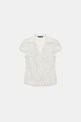 Camisa de cuello solapa y escote en pico, con mangas cortas y detalle de frunces centrales. Presenta cierre frontal con botones y estampado de lunares negros sobre fondo blanco.