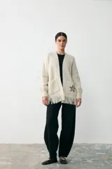 Chaqueta abierta de punto color crema, con diseño de corte recto, bolsillos frontales, flecos en el ruedo y detalle de estrella bordada en uno de los bolsillos.