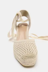 Alpargata de mujer con plataforma y taco cuña alto, confeccionada en rafia trenzada. La capellada es de tejido de punto calado color beige, con talón cerrado y tiras largas de tela para atar al tobillo.