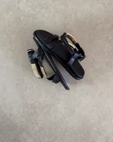 Sandalias planas de color negro con tiras finas y detalle decorativo de tubos metálicos dorados en el empeine. Cuentan con cierre de hebilla ajustable en el tobillo.