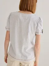 Remera de algodón con cuello redondo y mangas cortas con frunces en los hombros. Presenta detalles decorativos de cartera con botones en el lateral del cuerpo y en los puños, con terminaciones en lino.