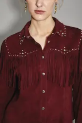 Camisa estilo western color bordó, confeccionada en gamuza caprina, con flecos largos en el canesú delantero y en las mangas. Presenta cuello con tachas metálicas plateadas.