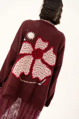 Saco de punto color vino con diseño holgado, cuello redondo y mangas largas. Presenta un gran bordado central en la espalda con forma de flor en tonos crema y rojo, acompañado de un pequeño bordado de sol y una frase bordada en la parte inferior.