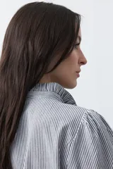 Camisa de rayas verticales con cuello mao, detalle de volados en el escote, bordados florales y recortes con calados. Posee mangas largas abullonadas con puños ajustados y cierre frontal con botones.