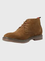 Bota tipo chukka de Hush Puppies, confeccionada en descarne color marrón, con cordones al tono, suela de goma y detalle de tirador en el talón con franjas rojas y negras.