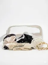 Kit de accesorios para el cabello que incluye un neceser transparente con borde blanco, dos scrunchies (uno negro con perlas y uno beige), dos clips livianos, una pinza dorada, un peine protector para desenredar y un peine para abrir ondas y rulos.