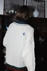 Buzo tejido de punto color terracota con diseño de rayas verticales en relieve. Presenta cuello en V con detalles de rayas en azul marino, blanco y negro, y puños acanalados a juego. Incluye un pequeño bordado en el pecho.