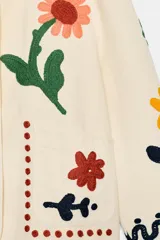 Chaqueta de algodón color crudo con cuello redondo, manga larga y bolsillos delanteros de plastrón. Presenta un diseño con bordados coloridos de flores y motivos abstractos a contraste, y cierre frontal con botones oscuros.