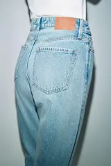 Jeans de denim azul claro lavado, tiro alto, corte Mom Fit con pierna recta y largo tobillero. Presenta cinco bolsillos y cierre frontal con cremallera y botón.