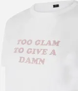 Remera de algodón blanca con cuello redondo y mangas cortas. Presenta una estampa frontal con la frase 'Too glam to give a damn' decorada con strass rosados y detalles de pliegues en los hombros.
