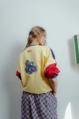 Buzo amarillo con mangas azul marino y puños rojos. Presenta un bordado de una flor en la espalda y un parche con el logo de la marca en el frente.