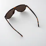 Lentes de sol con armazón de policarbonato negro y diseño de estilo aviador con puente superior plano. Las lentes son de color amarillo y cuentan con filtro UV400. Las patillas incorporan detalles metálicos dorados en las bisagras.