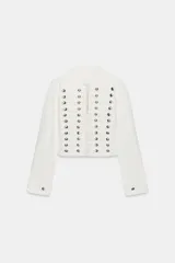 Chaqueta de denim blanca con cuello redondo, manga larga y corte entallado. Presenta un diseño de inspiración militar con múltiples tiras horizontales en el delantero decoradas con botones metálicos plateados, y cierre frontal con ganchos.