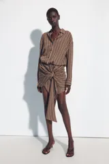 Vestido corto marrón con rayas verticales blancas, cuello camisero, manga larga y detalle de nudo y lazada en la falda. Cuenta con cierre frontal de botones.