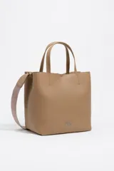 Bolso de hombro tipo tote bag de piel vacuna color beige, con doble asa de mano y correa ajustable de tejido. Presenta un cierre de cordón en la parte superior y un logo metálico en relieve en el frente. Incluye bolsillo interior con cierre de cremallera.
