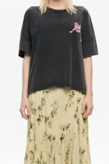 Camiseta de algodón color blanco hueso, de corte boxy y holgado, con escote redondo y mangas cortas. Presenta un pequeño logo en relieve de color rojo en el área del pecho.