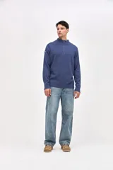 Buzo de punto color azul, con cuello alto y cierre frontal parcial. Presenta un diseño con textura acanalada en los hombros y mangas, y un pequeño bordado en la manga derecha.