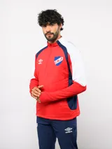 Buzo de entrenamiento Umbro del Club Nacional de Football, con cierre frontal parcial y cuello alto. Presenta un diseño con bloques de color azul, blanco y rojo, con panel frontal acolchado y mangas largas con aberturas para pulgares.