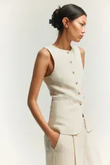 Chaleco largo color blanco hueso, con cuello redondo, cierre frontal con botones dorados, bolsillos delanteros ribeteados y abertura en la parte posterior. Forrado.