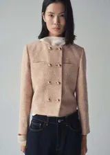 Chaqueta de tweed en color beige con diseño recto, cuello redondo y cierre frontal de doble botonadura con botones dorados. Presenta dos bolsillos de parche en el pecho y forro interior.