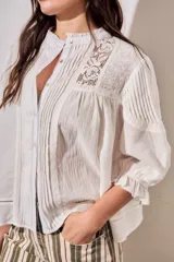 Blusa de algodón peinado color off-white, con cuello mao, cierre frontal de botones, mangas tres cuartos abullonadas con puños con volados, detalles de alforzas y apliques de encaje en el frente y los hombros.