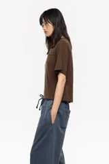 Camiseta de manga corta y escote redondo, color beige claro, con corte boxy. Presenta una hilera de tachuelas metálicas negras decorativas en la parte superior del pecho y en las costuras de los hombros.