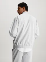 Campera deportiva Calvin Klein color gris claro, de sarga de satén brillante, con cuello alto, mangas raglán, puños y bajo elásticos, cierre de cremallera, bolsillo con cremallera en el pecho, bolsillos con botón de presión en la cintura, forro de malla transpirable y logo monograma estampado en el pecho.