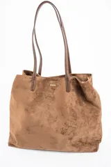 Bolso tipo tote de gran tamaño confeccionado en gamuza sintética color tostado. Presenta un diseño minimalista con asas largas de hombro y una pequeña placa metálica frontal con el logo de la marca.