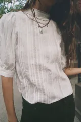 Blusa blanca de cuello alto con detalles de puntilla, manga corta abullonada y cierre posterior con botón.